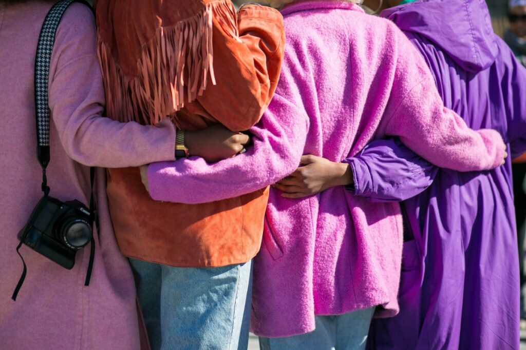 Four people, wearing colorful jackets, stand in a row with their arms around each others backs. One person has a camera hanging by their side.