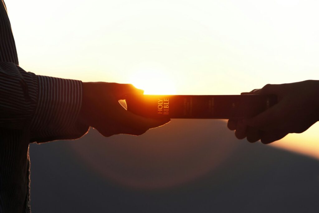 Two hands exchanging a book with a sunset in the background.