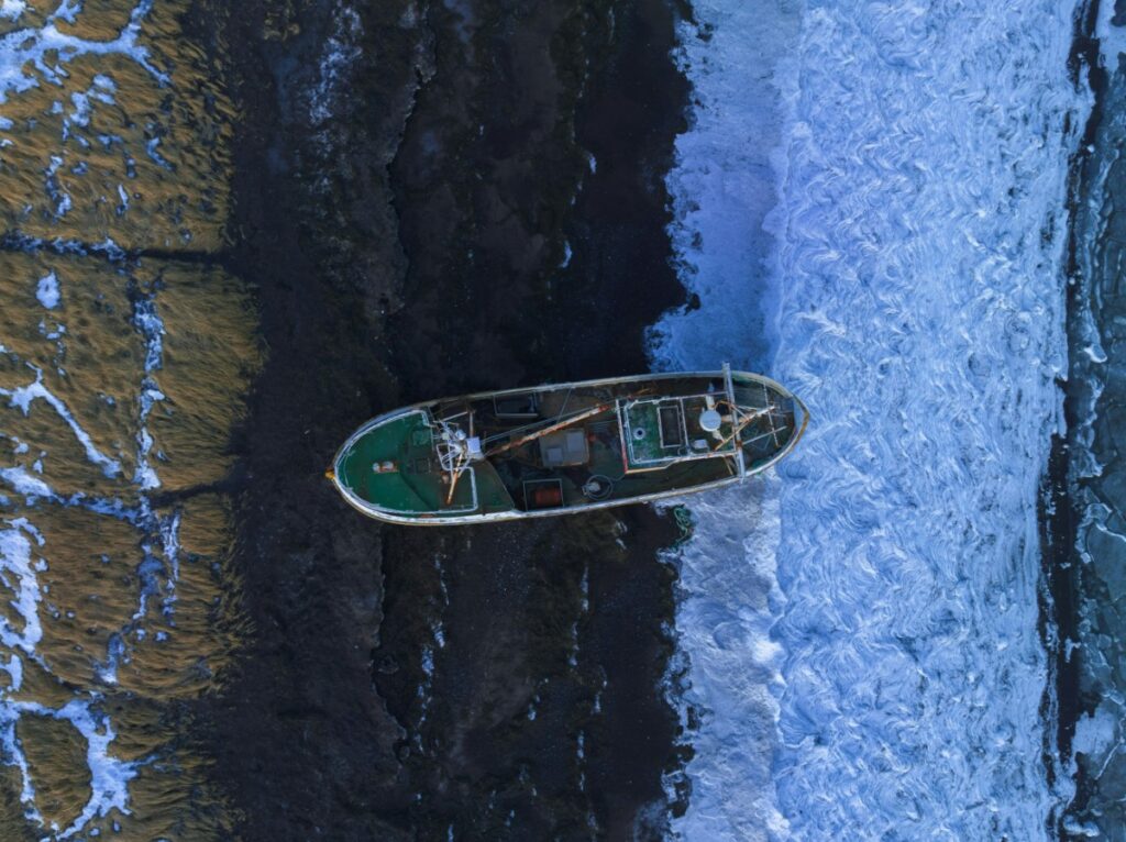 Aerial view of a shipwrecked boat half-submerged at the shoreline, with waves crashing on one side and exposed seabed on the other.