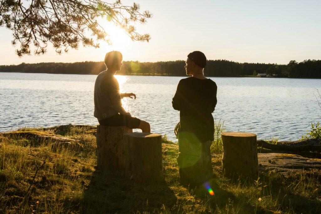 Two people sit on tree stumps by a lake at sunset, engaged in conversation.