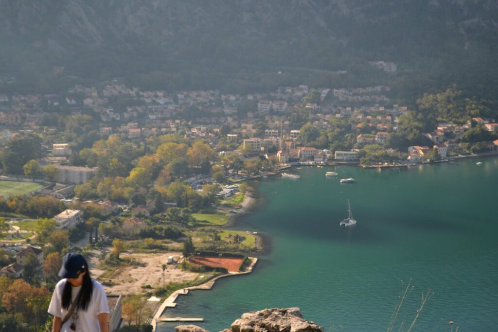 A person stands with a view of a coastal town featuring houses, trees, and a sailboat on the water, with mountains in the background.