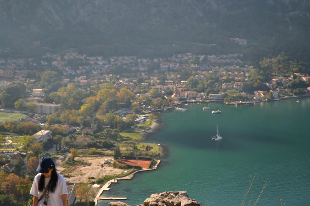 A person stands with a view of a coastal town featuring houses, trees, and a sailboat on the water, with mountains in the background.