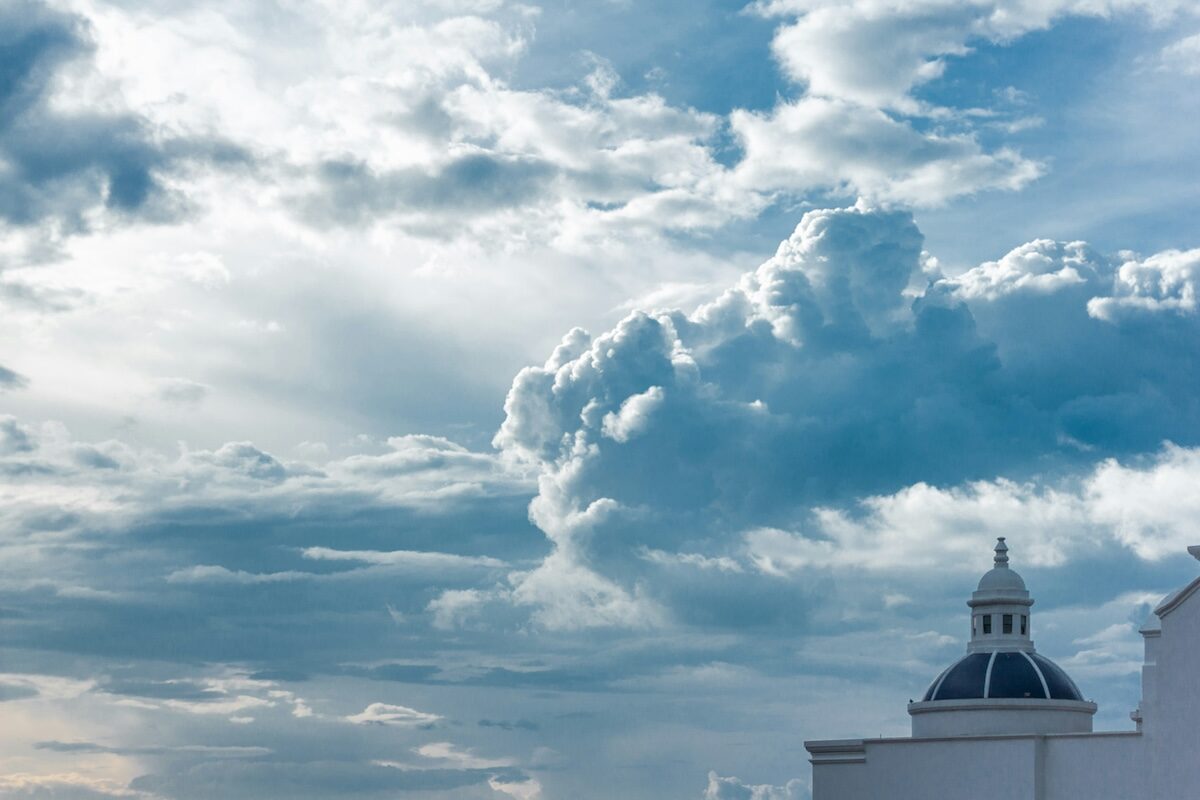 A white building with a domed roof stands beneath a sky filled with large, dramatic clouds.