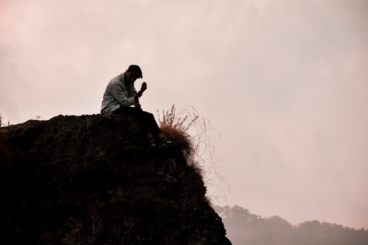 A person sits alone on the edge of a rocky cliff, looking down with their hand on their face against a cloudy sky backdrop.