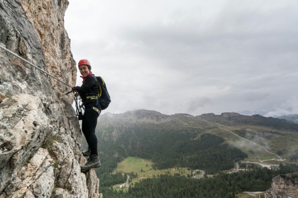 Person in climbing gear ascends a rocky cliff face with a forested landscape in the background.