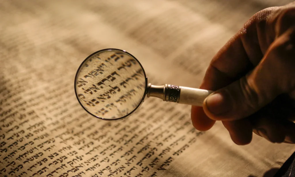 A hand holds a magnifying glass over a page with Hebrew text, enlarging the characters under the lens.