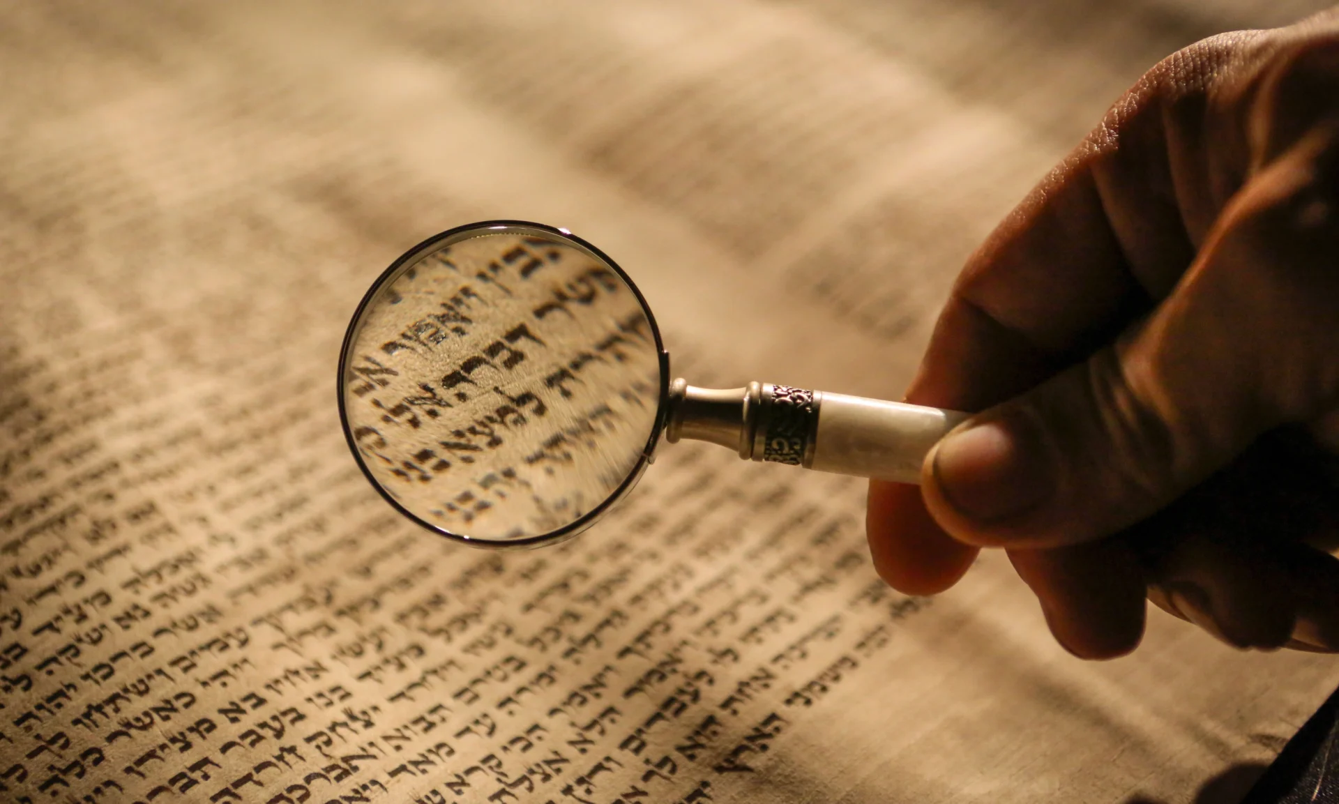 A hand holds a magnifying glass over a page with Hebrew text, enlarging the characters under the lens.