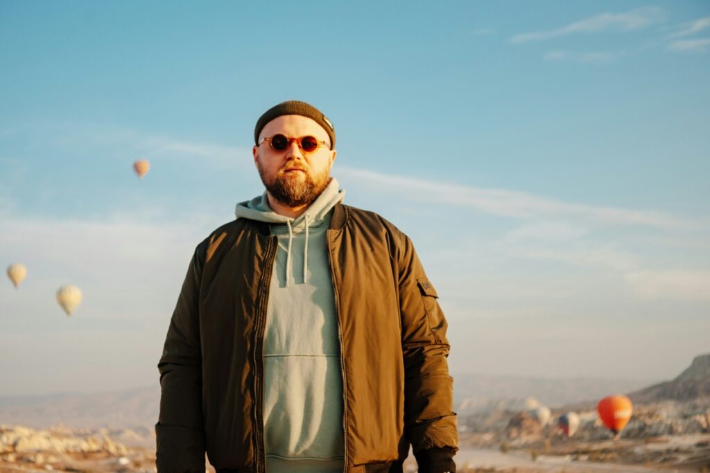 A person wearing a beanie and sunglasses stands outdoors, with hot air balloons visible in the background against a clear sky.