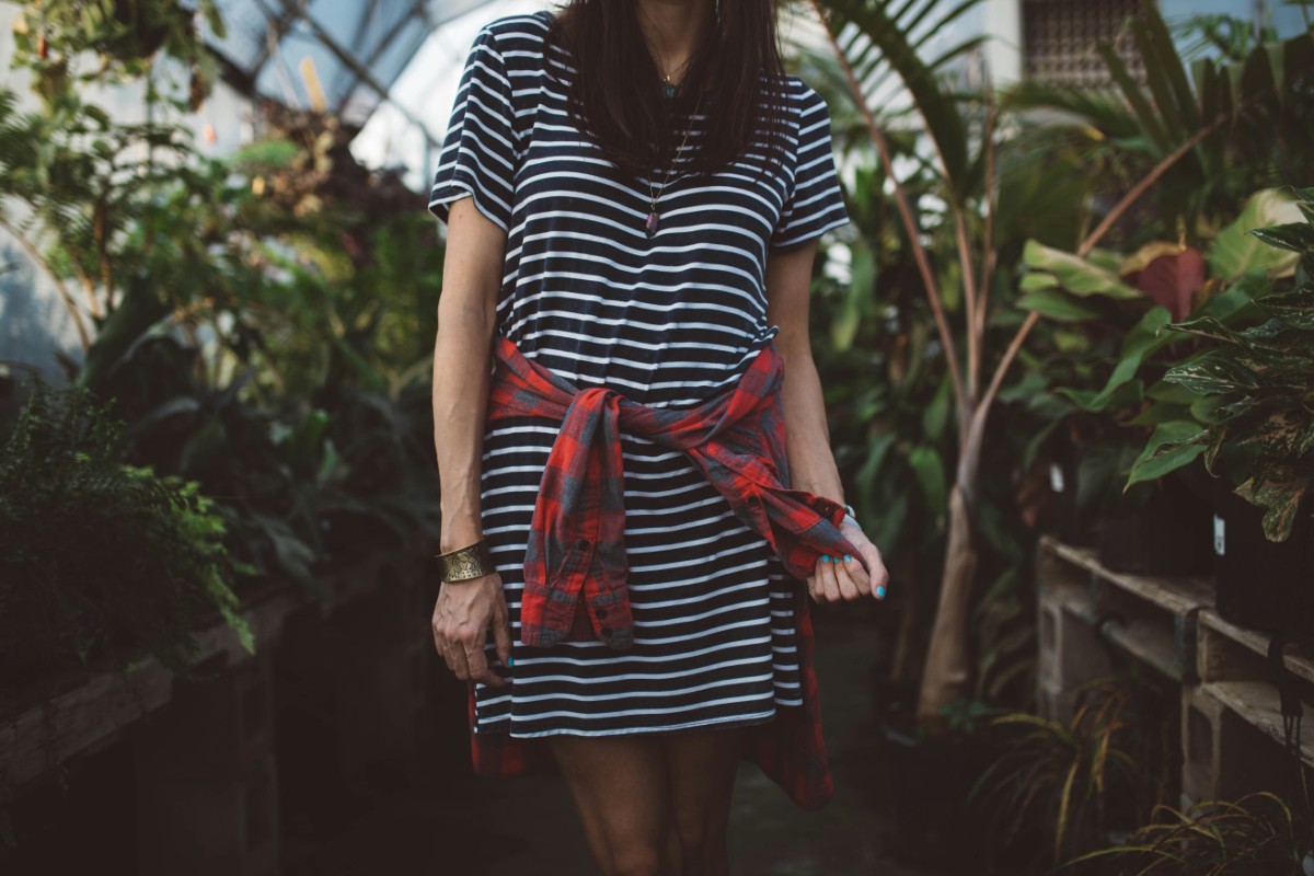 A person in a striped dress stands in a garden. A red plaid shirt is tied around their waist. Green plants surround them.