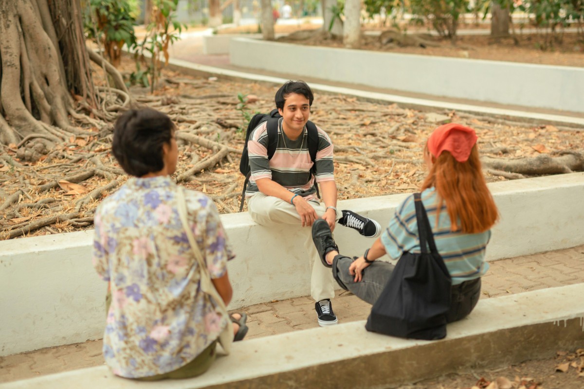Three people sitting on a low wall outdoors, engaged in conversation. One person has a backpack, another has a red hair accessory. Trees and a path are in the background.