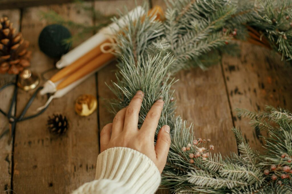 A hand touching a pine wreath on a wooden surface, surrounded by candles and pinecones.