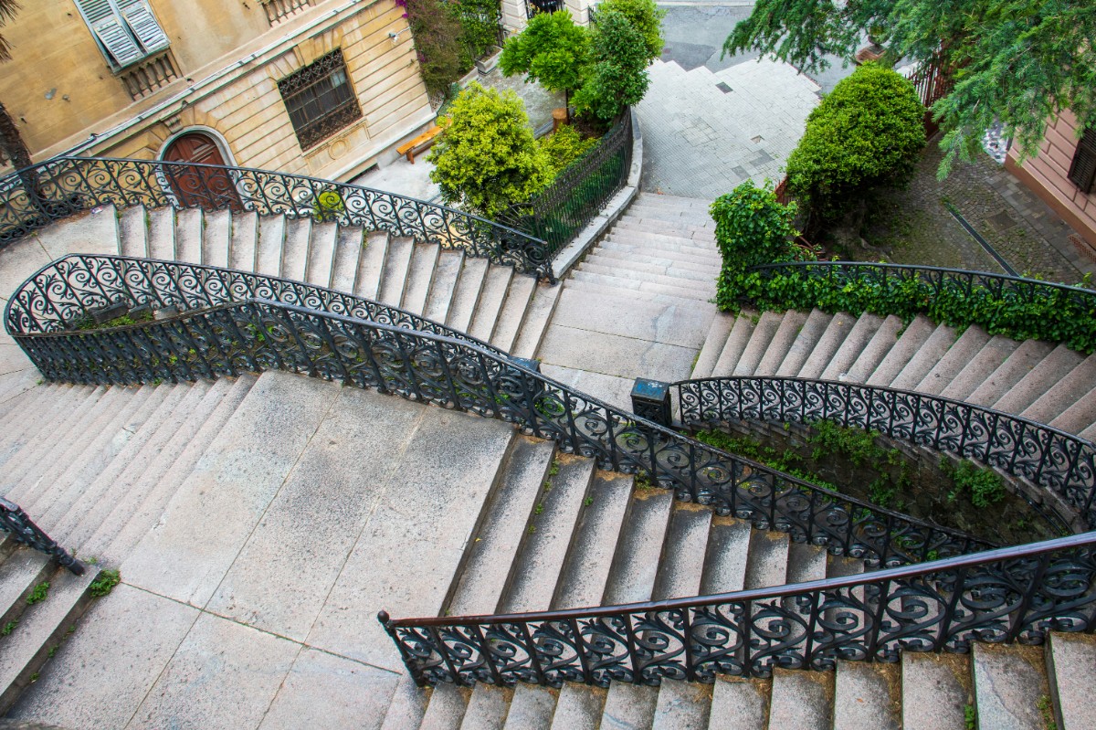 A stone staircase with decorative iron railings splits into two sections, leading down to a garden path lined with green shrubs and trees.