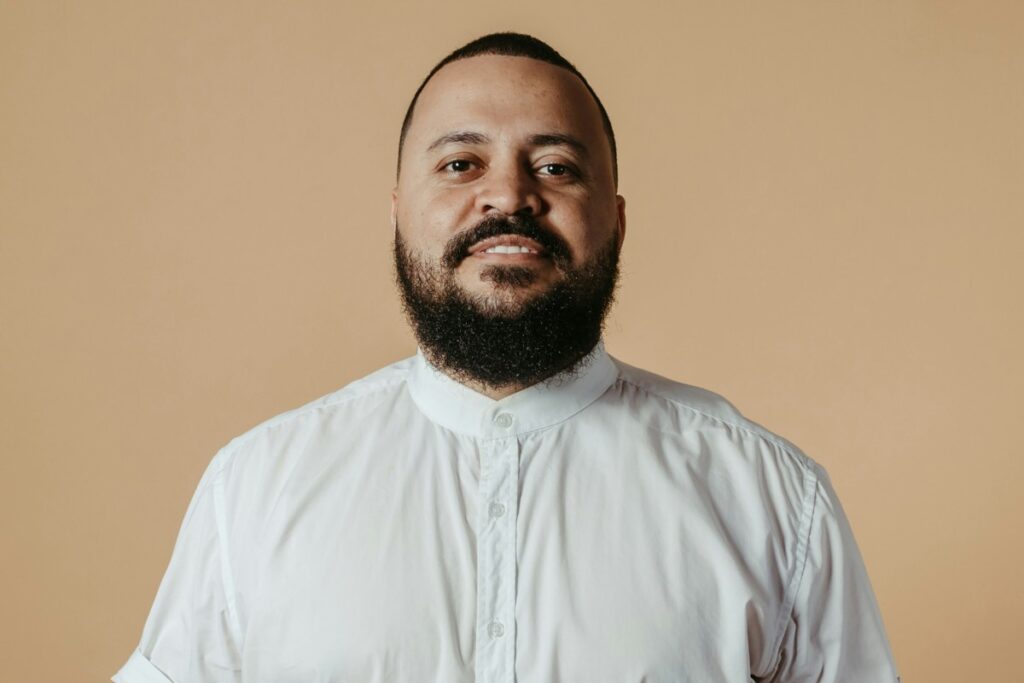 Man with a beard and short hair wearing a white button-up shirt, standing in front of a beige background.