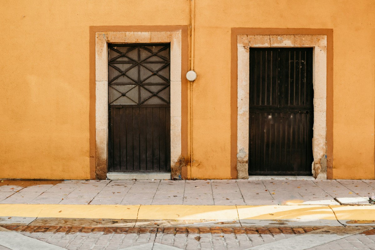 Two black doors set in an orange wall, with a yellow stripe at the bottom, over a tiled sidewalk.