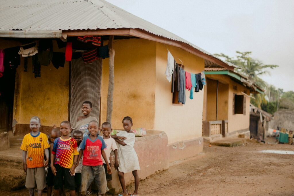 Children stand in front of a house with a metal roof. Clothes are hanging to dry on a line next to the building. The ground is muddy, and another structure is visible in the background.
