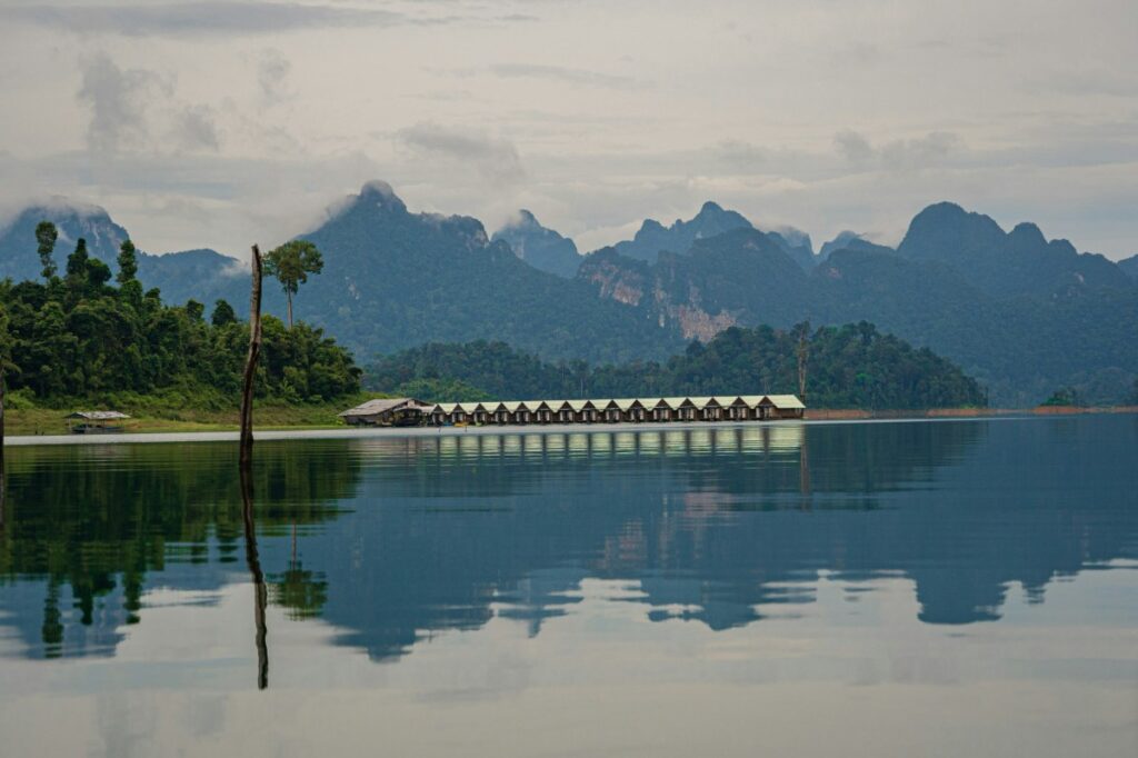Row of cabins on a calm lake with forested shoreline and mountainous background under a cloudy sky.