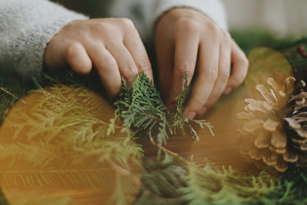 Hands arranging green foliage and a pine cone on a surface, with a decorative focus.