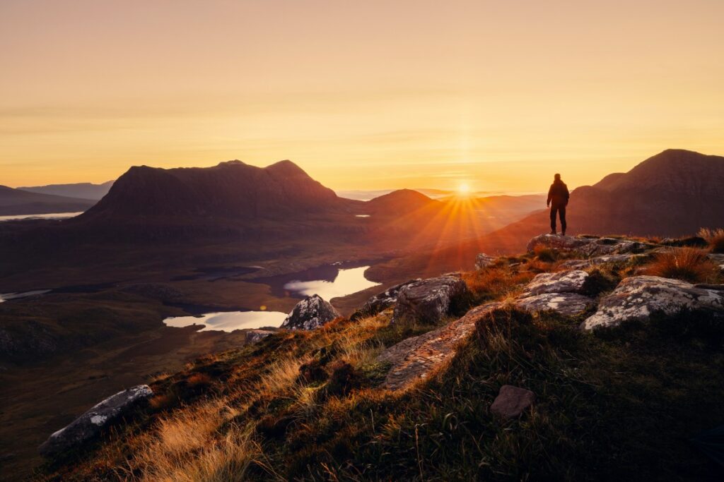 A person stands on a rocky hilltop, overlooking a landscape of hills and lakes at sunrise.
