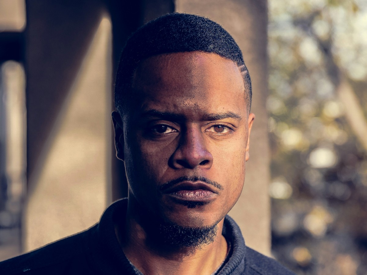 Man with short hair and facial hair, standing outdoors in natural light, with a serious expression. Blurred background with foliage.