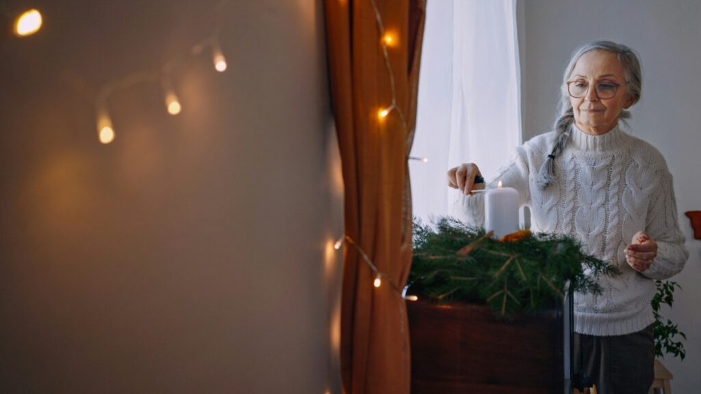 Older woman in a cozy sweater arranges candles on a decorated table with pine branches. Warm string lights hang nearby, adding a festive ambiance.