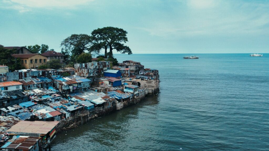 A cluster of coastal shanty houses extends into the sea under a blue sky. Large trees and distant boats are visible.