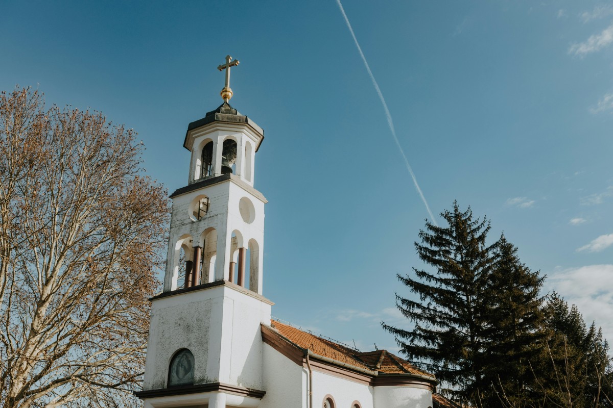White church with a tall bell tower and a cross on top, set against a blue sky with a visible contrail. Trees are on either side.
