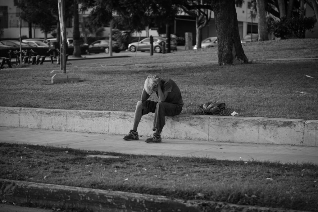 A person sits alone on a low concrete wall in a park, leaning forward with their head down, surrounded by trees and parked cars in the background. Black and white image.