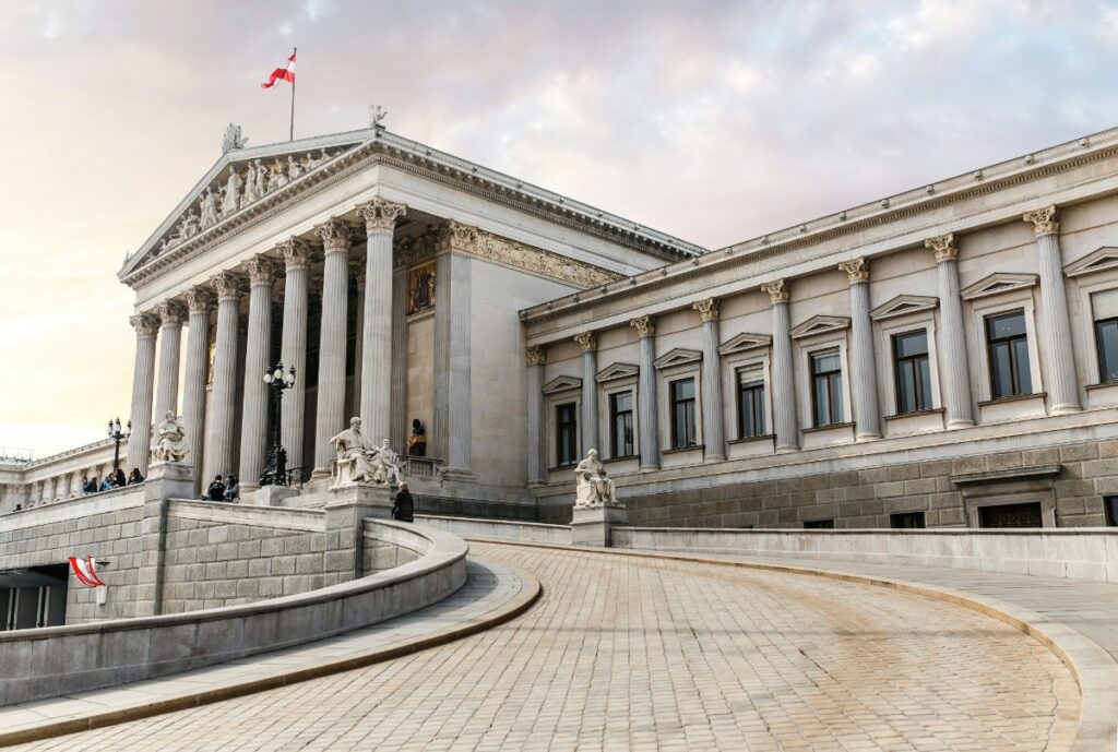 Neoclassical building with columns and statues, featuring a curved ramp and a flag on top, under a cloudy sky.