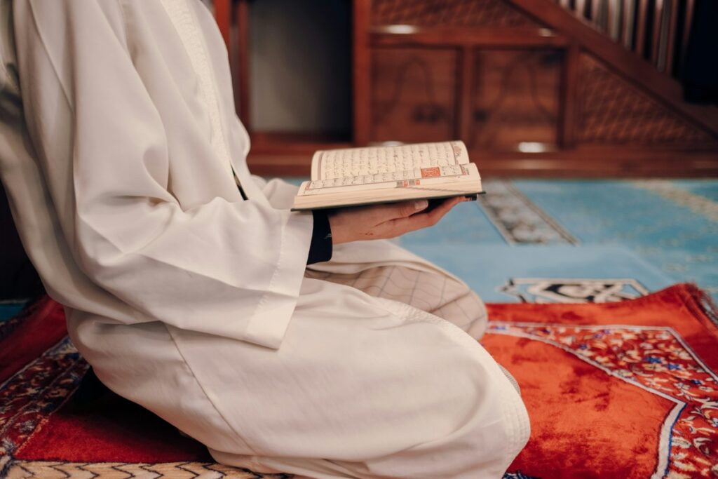 Person in a white garment kneeling on a red and orange patterned rug, holding an open book.