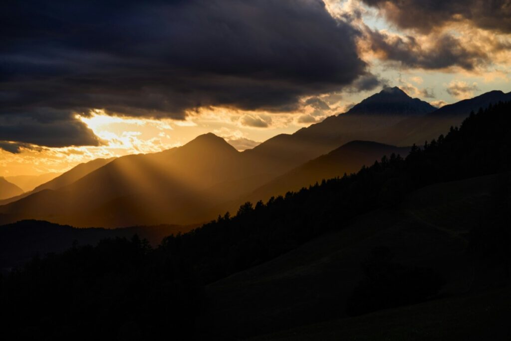 Sunset over mountains with dramatic clouds and sun rays piercing through, casting a warm glow on the landscape.