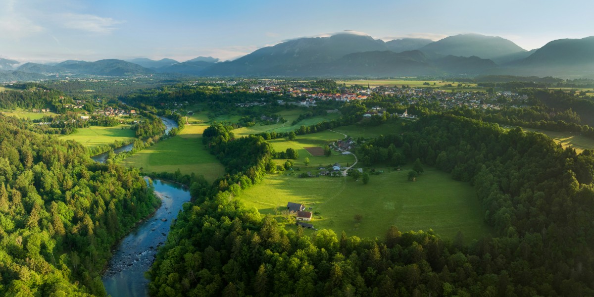 Aerial view of a lush, green valley with a river winding through fields and forests. Mountains rise in the background, and a small town is visible under a clear blue sky.
