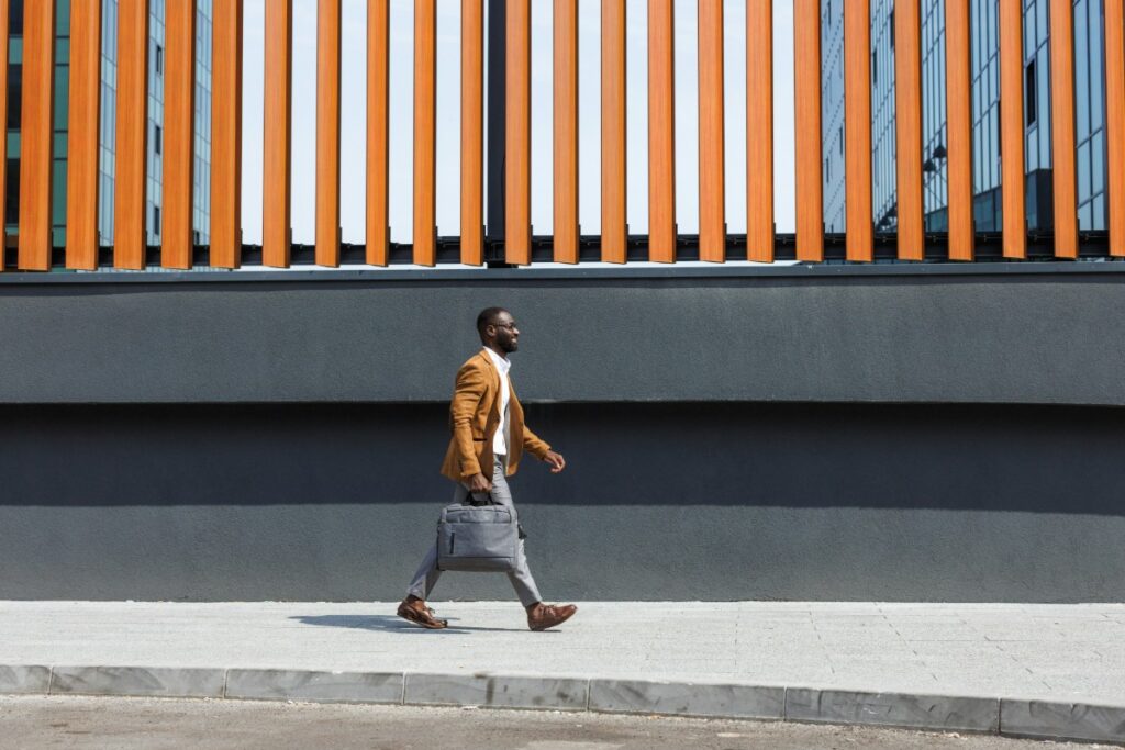 Man walking on the sidewalk, wearing a brown jacket and carrying a briefcase, in front of a modern building with vertical orange and gray stripes.