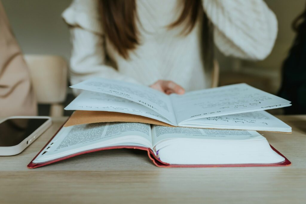 A person leafs through a book while another lies open on a table, next to a smartphone.