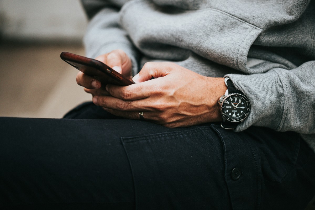 Person in a gray sweatshirt and dark pants holding a smartphone. A large black watch is visible on their wrist.