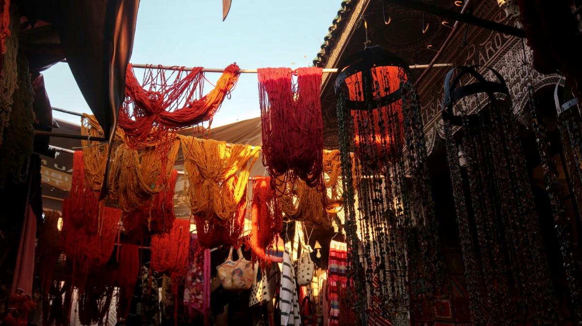 Colorful textiles and beads hang in a dimly lit market, casting shadows.