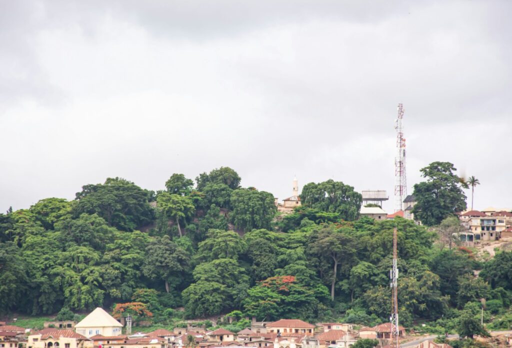 A hilly landscape with dense green trees and scattered houses. A tall communication tower is visible, and the sky is overcast.