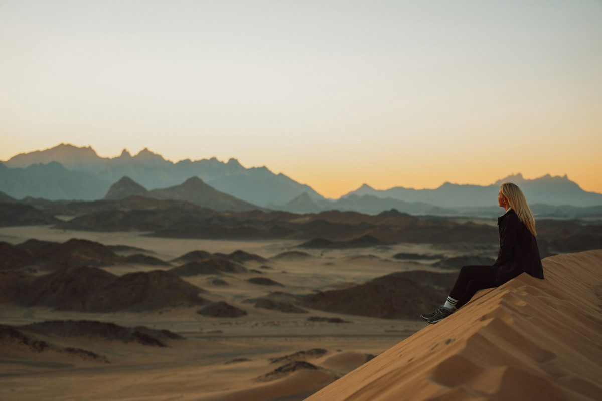 Person sitting on a sand dune, overlooking a vast desert landscape with distant mountains under a sunrise or sunset sky.