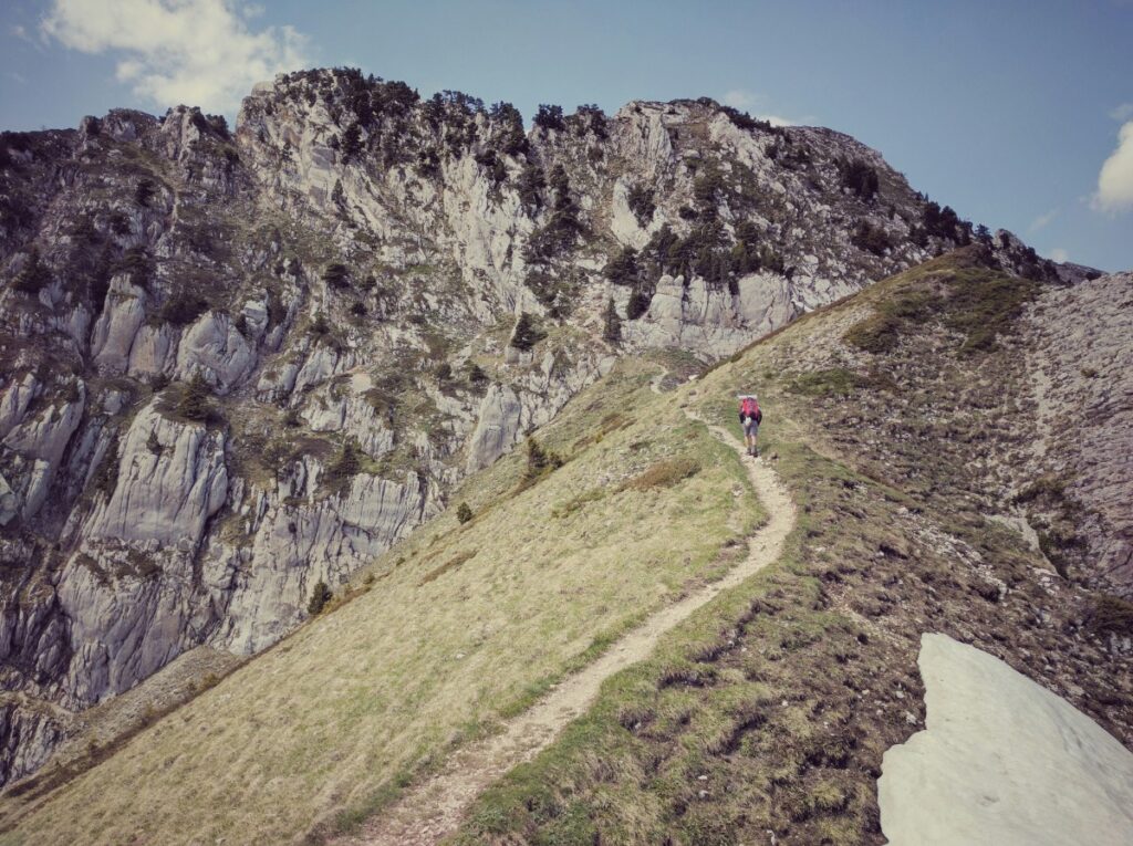 Narrow hiking trail on a mountain ridge with rocky cliffside; a person walks in the distance under a clear blue sky.