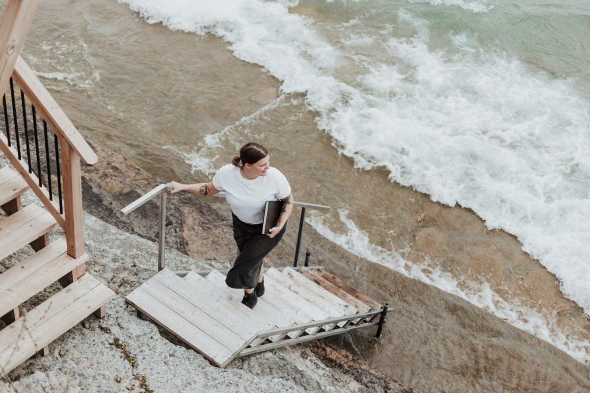 Person in a white shirt holding a laptop, walking up wooden stairs by the ocean, with waves crashing nearby.