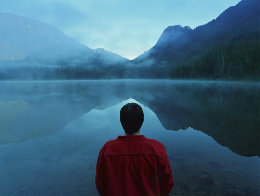 Person in a red jacket stands by a calm lake, gazing at fog-covered mountains in the background.