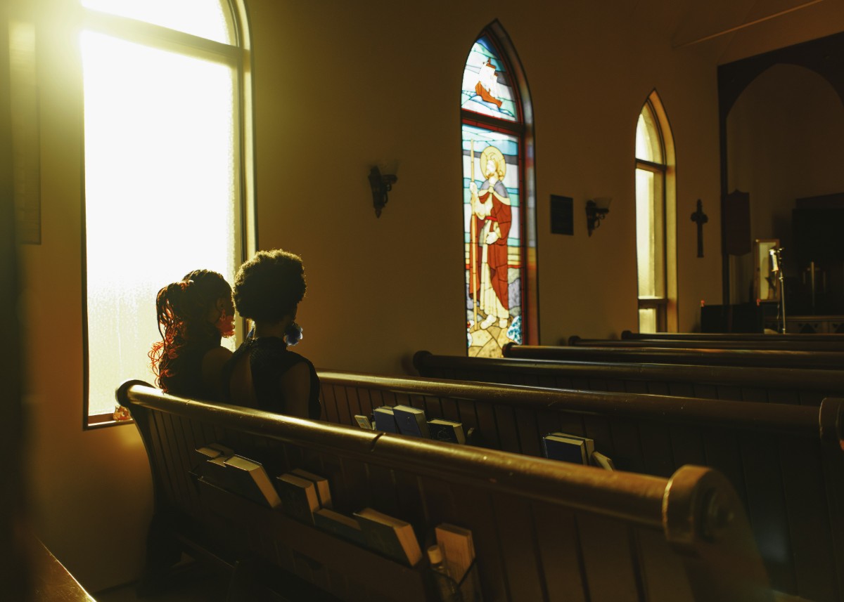Two people sitting on a wooden pew in a dimly lit church, with sunlight streaming through stained glass windows.