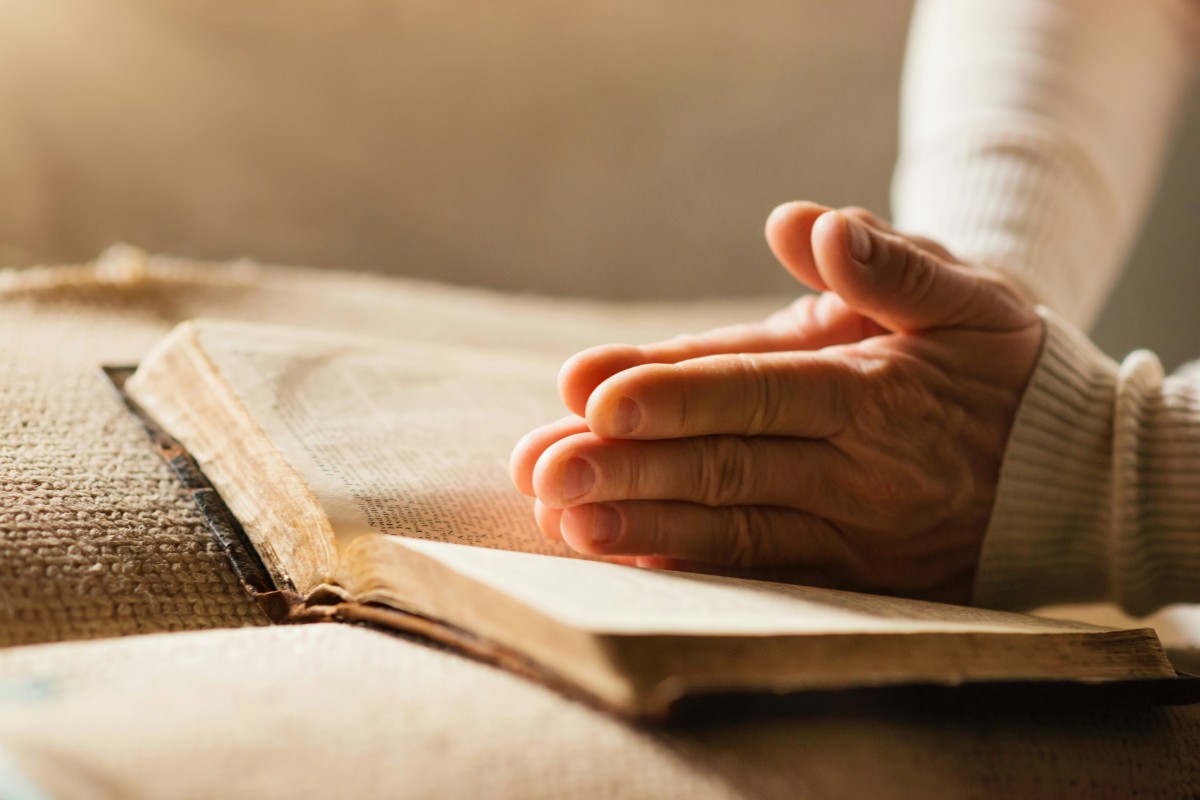 A person holds their hand over an open book on a burlap surface, with soft lighting filtering in from the side.