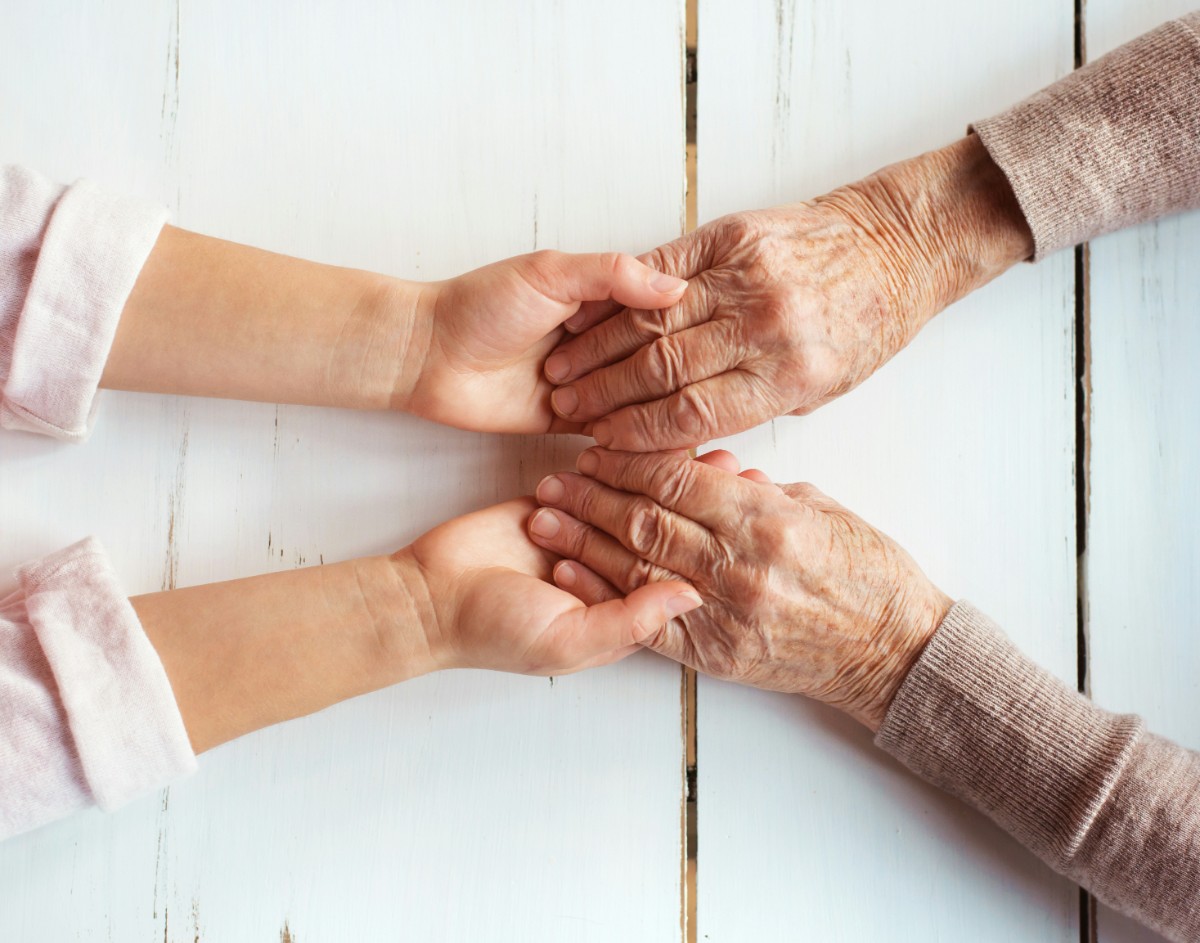 Two pairs of hands, one younger and one older, gently holding each other on a white wooden surface.