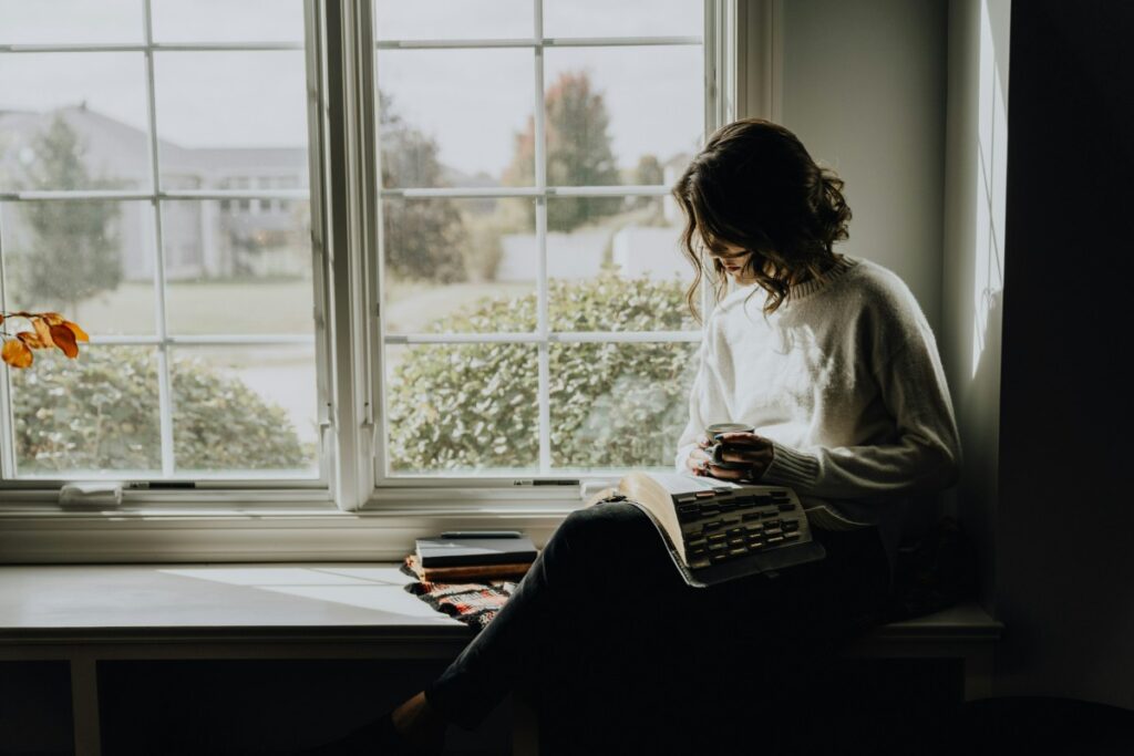 Person sitting by a window, reading a book. Dim natural light illuminates the scene.