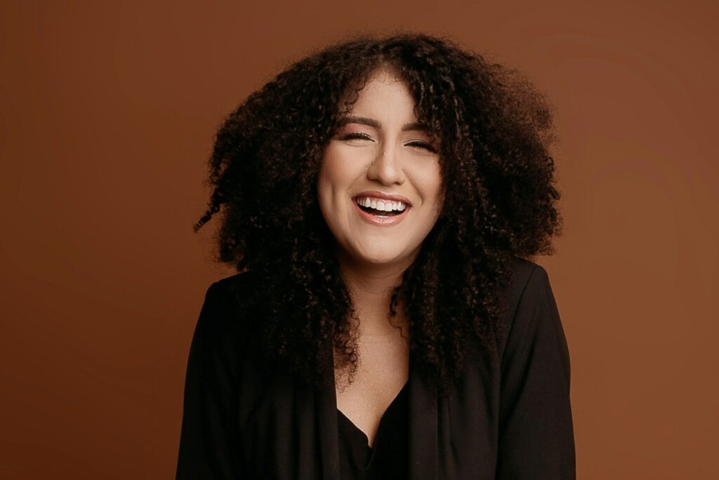 Smiling person with curly hair against a brown background, wearing a black top.