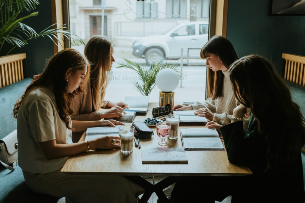 Four women sitting at a table in a cafe, reading menus. A car is visible outside the window.