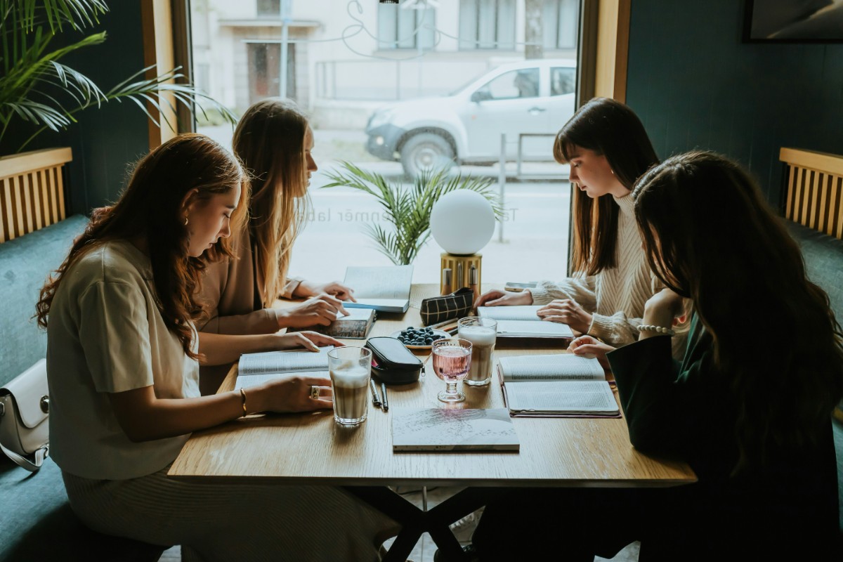 Four women sitting at a table in a cafe, reading menus. A car is visible outside the window.