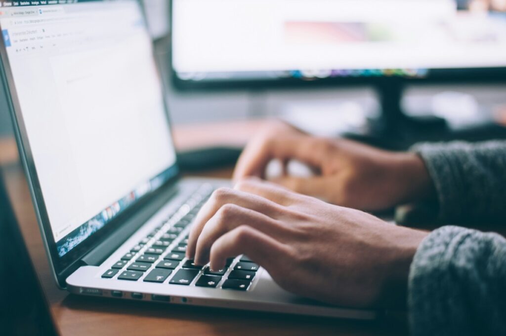 Hands typing on a laptop keyboard with a blurred monitor in the background.