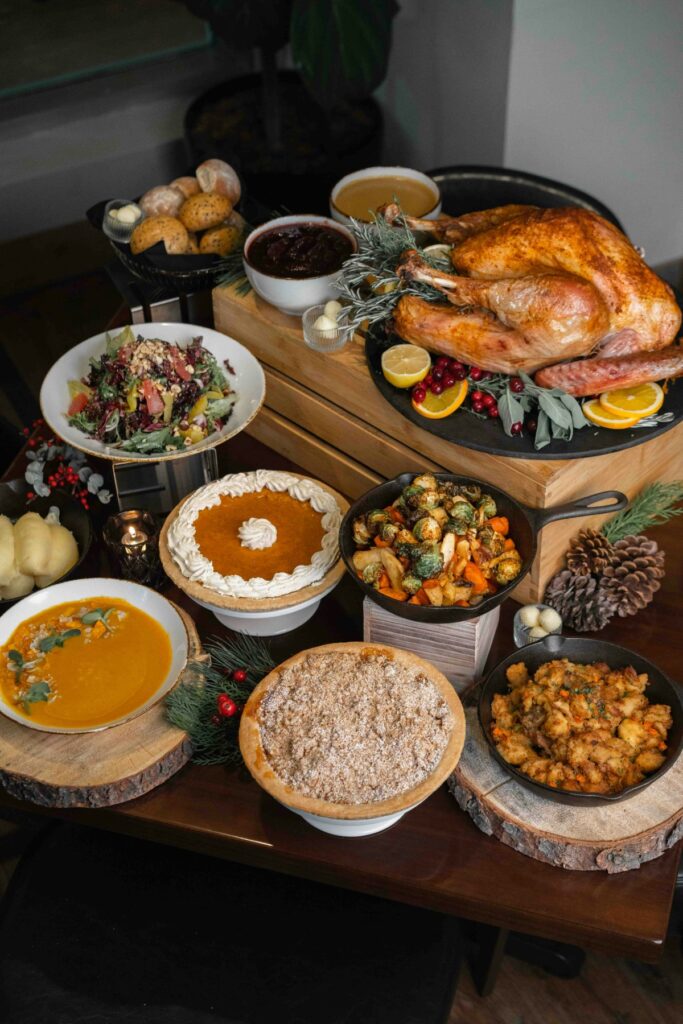 A table with a roasted turkey, various side dishes including salads, roasted vegetables, and bowls of sauce, alongside a pumpkin pie, muffins, and assorted fruits.