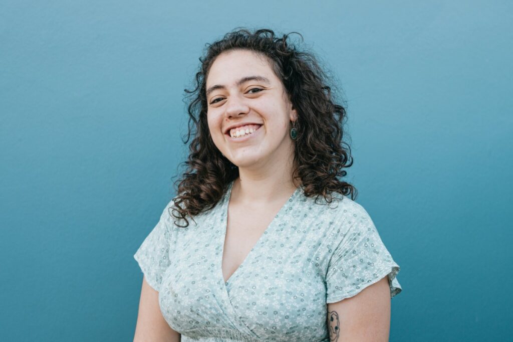 Person with curly hair smiling in front of a blue background, wearing a light floral dress.